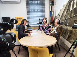 Trzy osoby w słuchawkach siedzą przy drewnianym stole z mikrofonami, pogrążone w dyskusji podcastowej. Kamery i sprzęt nagrywający misternie uchwycają tę bogatą w kulturę konfigurację.