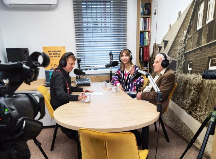 Trzy osoby w słuchawkach siedzą przy drewnianym stole z mikrofonami, pogrążone w dyskusji podcastowej. Kamery i sprzęt nagrywający misternie uchwycają tę bogatą w kulturę konfigurację.