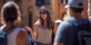 Kobieta w okularach przeciwsłonecznych i kapeluszu z szerokim rondem rozmawia z ludźmi podczas słonecznej wycieczki szkolnej w Warszawie.